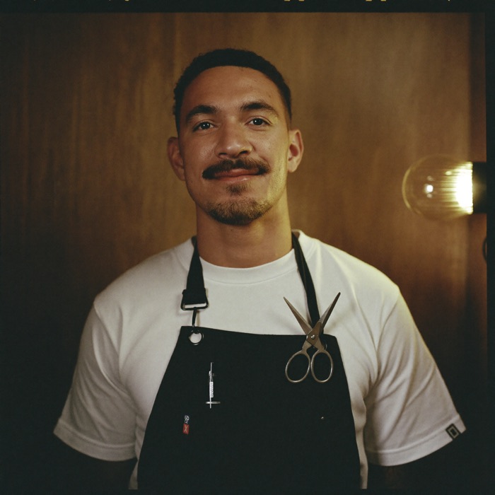 A barber in a black apron holding clippers in front of a warm oak wall.