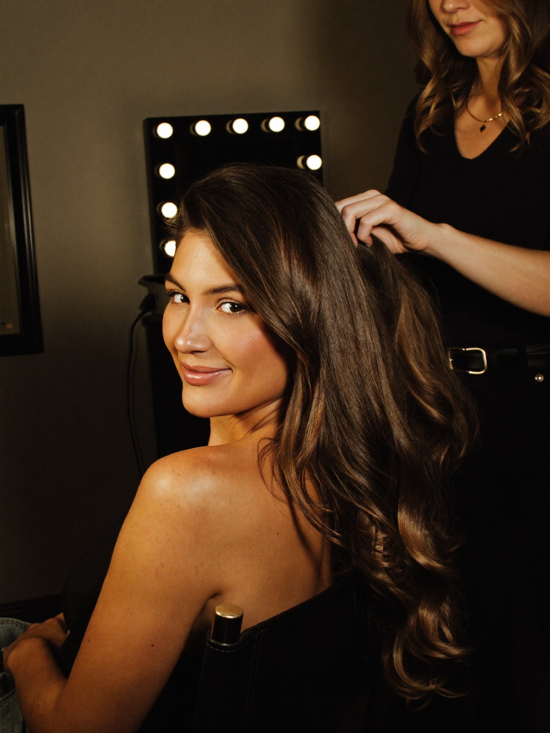 A client turning in the chair with freshly finished long hair while her stylist lifts a last section, at a bulb-lit mirror.