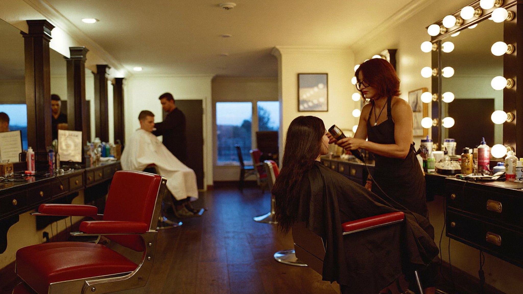 The C&C floor at dusk: a stylist blow-drying a client at a bulb-lit mirror, with the row of red barber chairs and a barber mid-cut behind her.