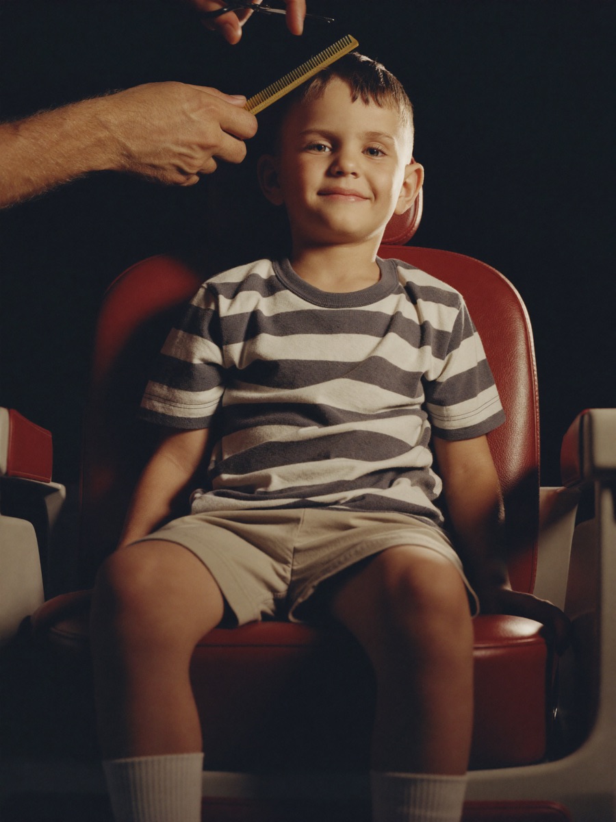 A young boy on a booster seat in a red barber chair, mid haircut.